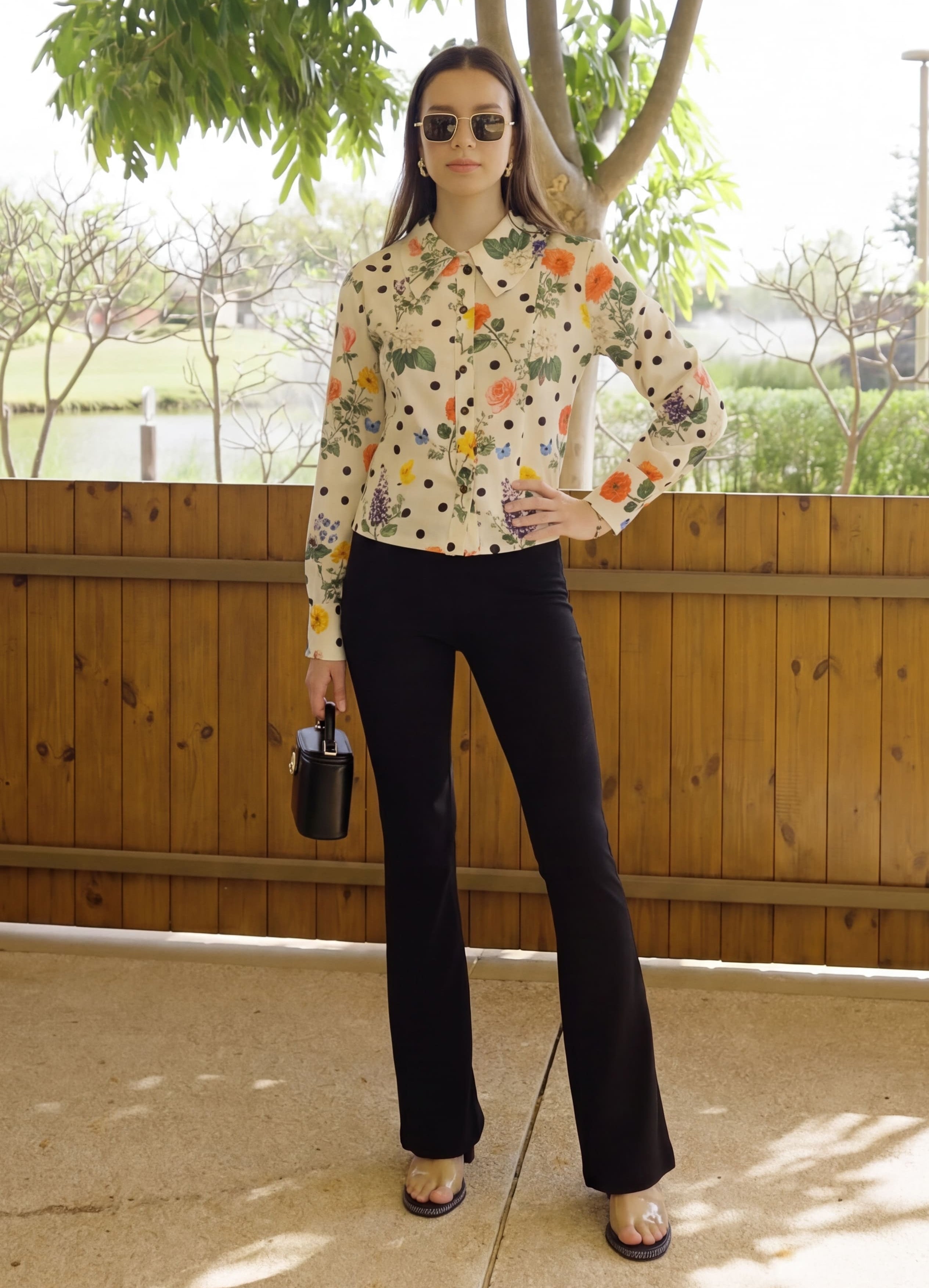 Woman in a patterned blouse and black pants standing under a wooden pergola with trees in the background.
