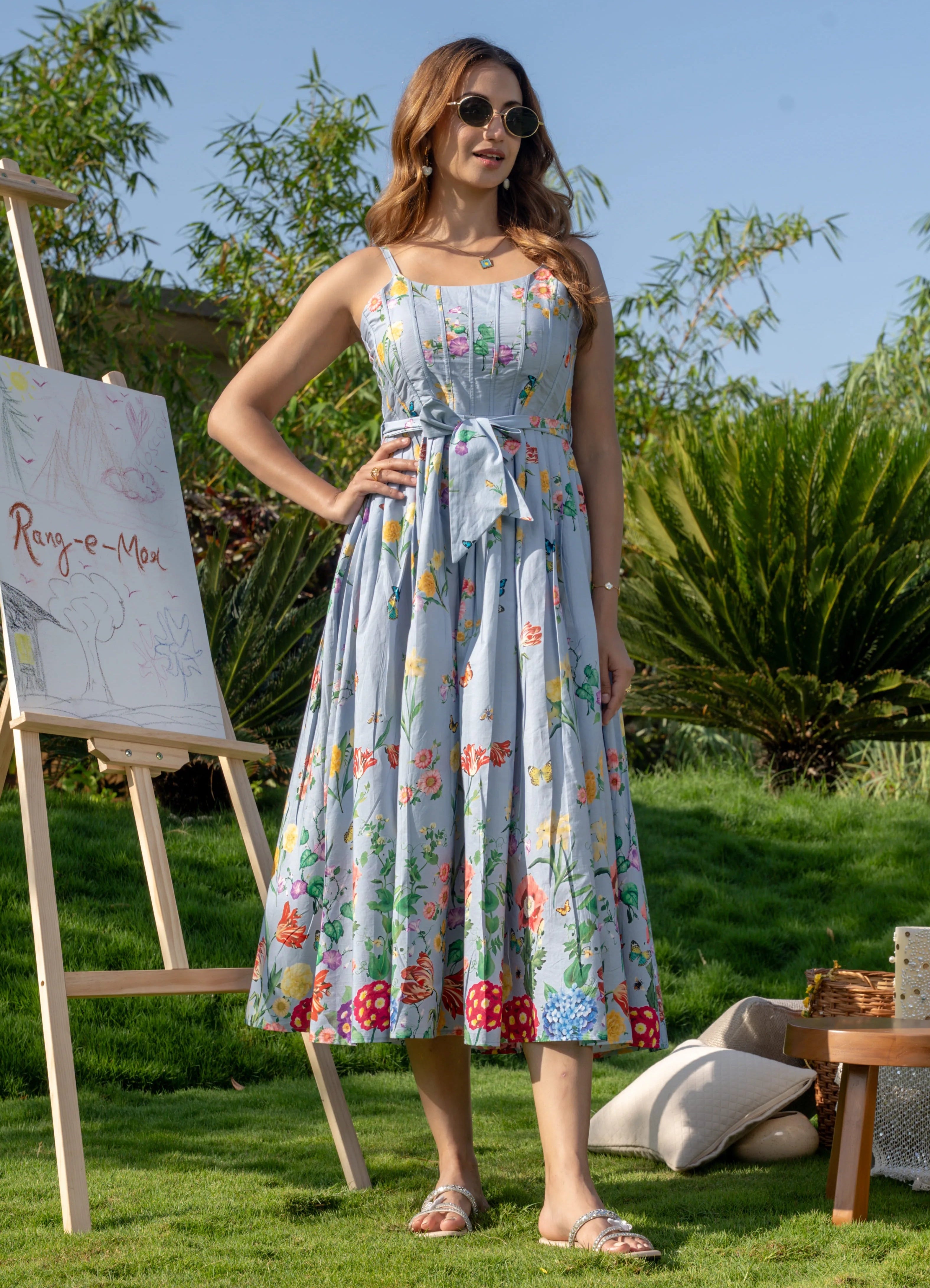 Woman in a floral dress standing outdoors with a painting easel and art supplies.