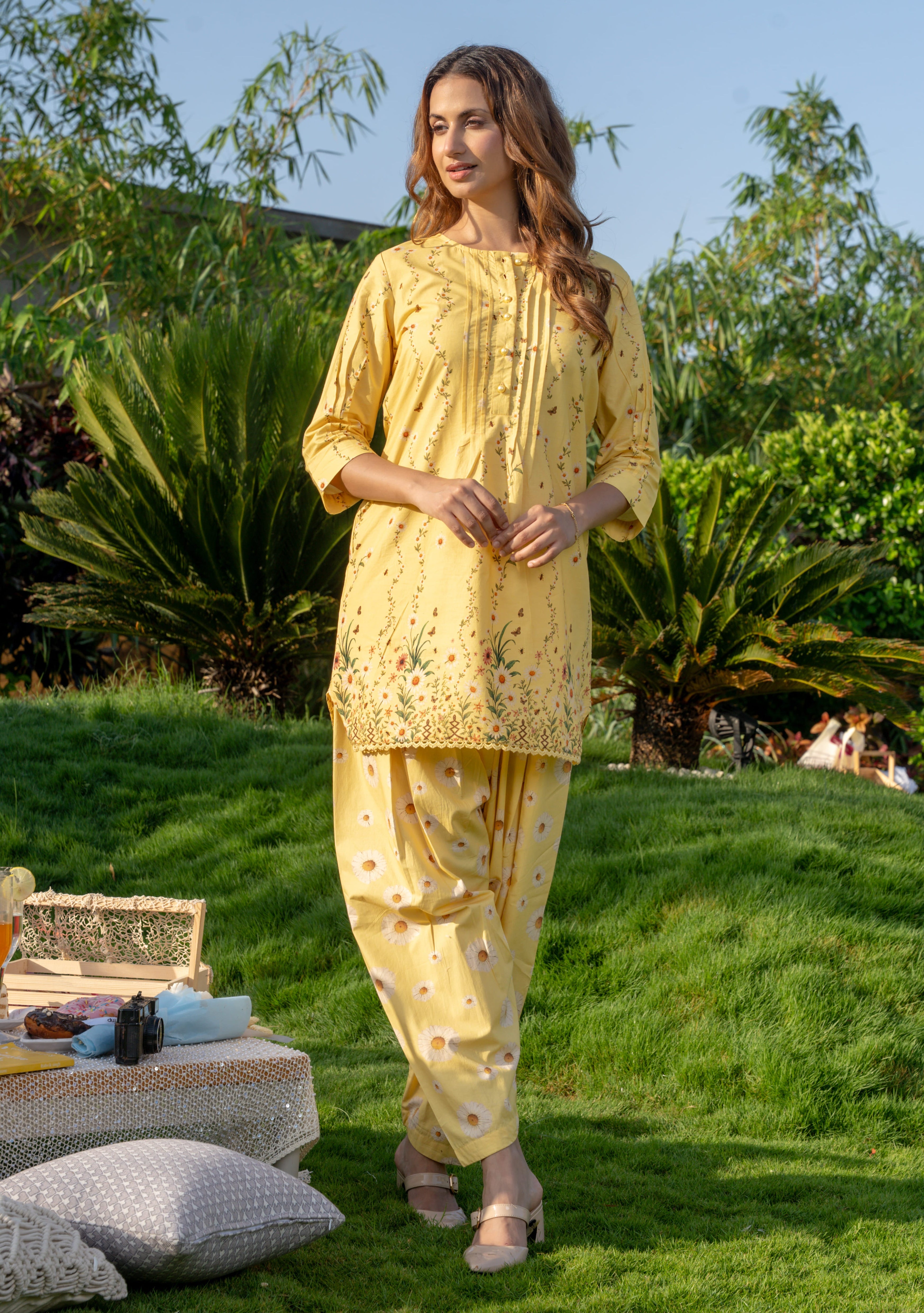 Woman in a yellow traditional outfit standing outdoors on a grassy area with greenery.