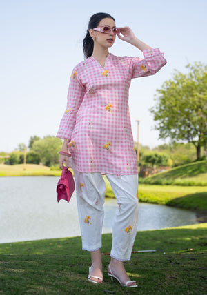 Woman in a pink checkered outfit with white pants standing by a pond with greenery in the background