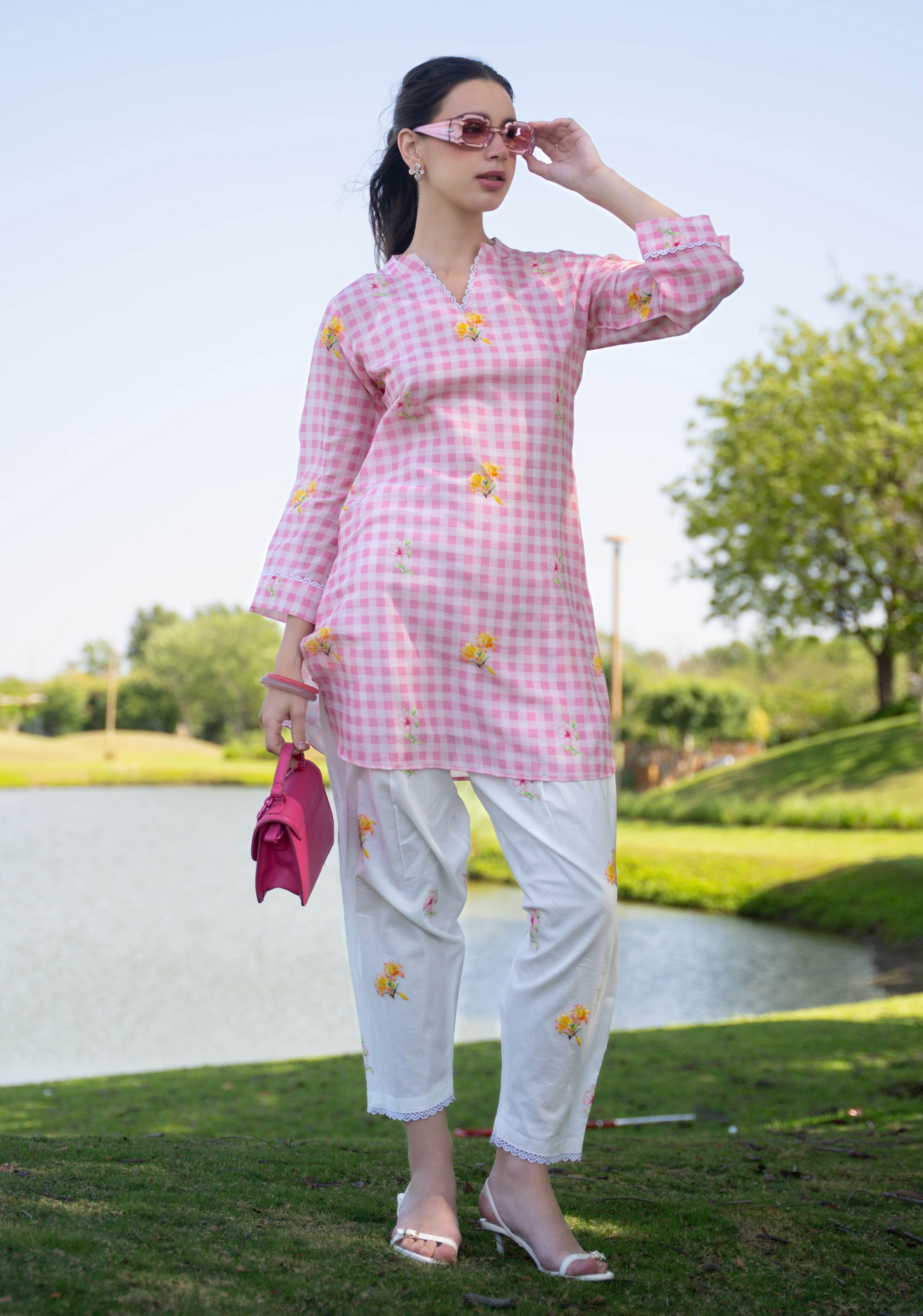 Woman in a pink checkered outfit with white pants standing by a pond with greenery in the background