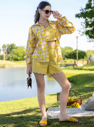 Woman in yellow checkered shirt and shorts standing by a lake with picnic blanket and basket.