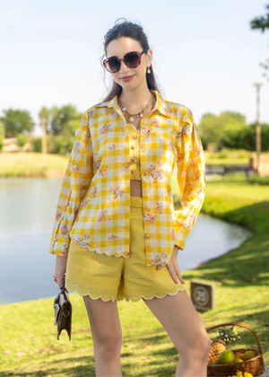 Woman in a yellow checkered shirt and shorts standing by a pond with a scenic background.