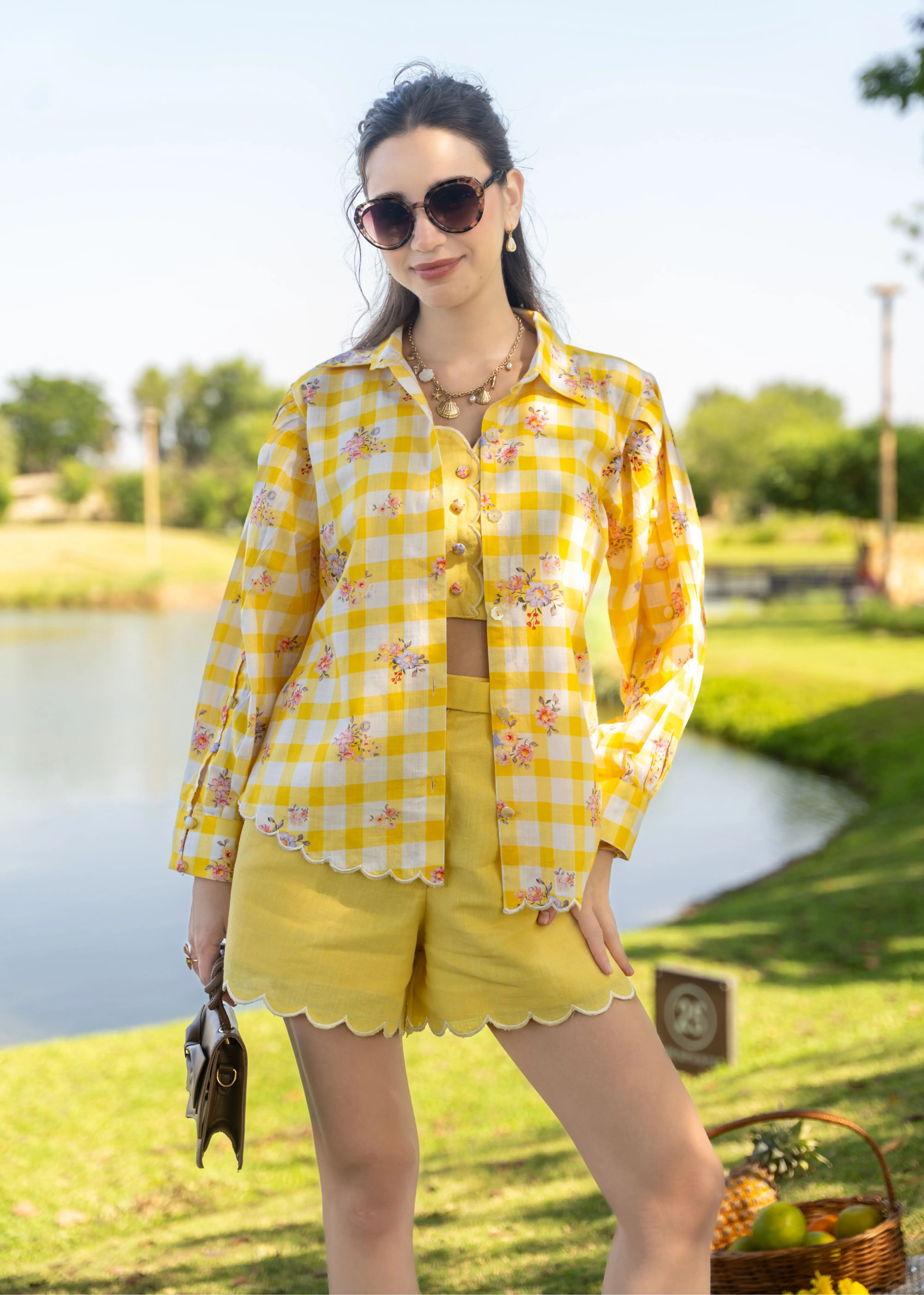 Woman in a yellow checkered shirt and shorts standing by a pond with a scenic background.